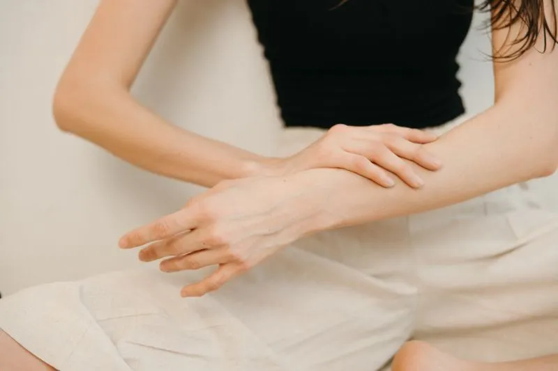 Woman in a black top and beige linen trousers massaging her forearm with the flat of her hand