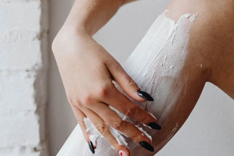 A hand with black-painted nails spreading a thick layer of white cream over a leg, in front of a white brick wall