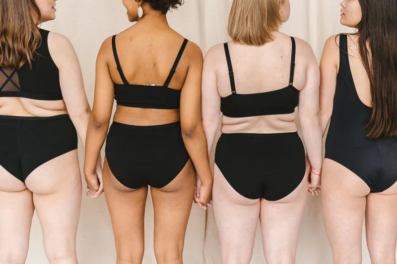 Four women seen from behind in black swimwear and underwear, with different body shapes and skin tones, holding hands