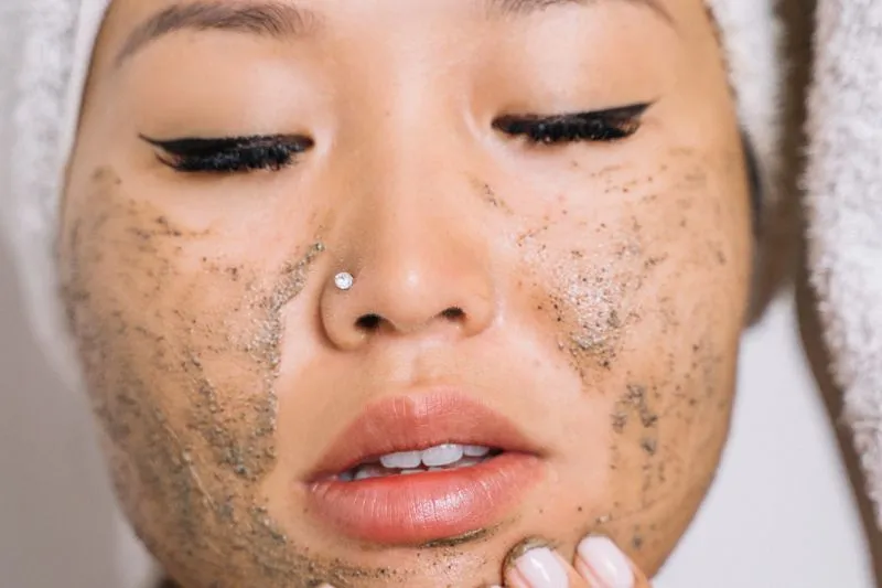 Close-up of a face, eyes closed, covered in a grainy scrub, hair wrapped in a towel