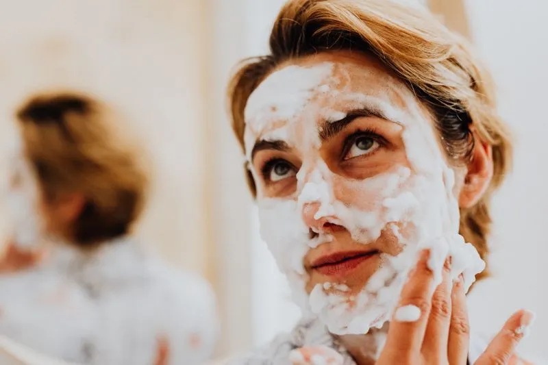 A woman with cleansing foam across her face, working it in with her fingertips in front of a mirror