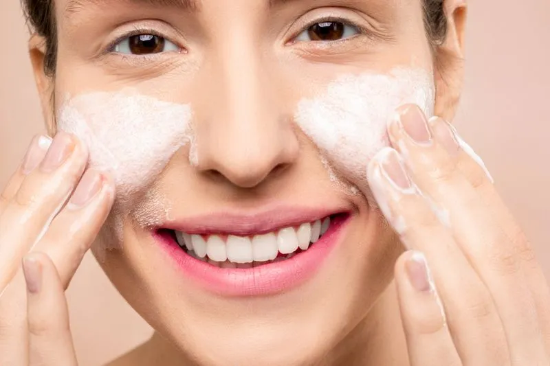 A smiling woman spreading white foaming cleanser across her cheeks with her fingertips