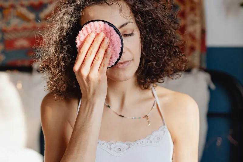 A curly-haired woman holding a pink reusable make-up remover pad over one closed eye