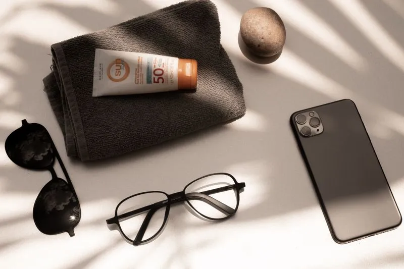 SPF50 sunscreen tube on a towel, with sunglasses, reading glasses, a pebble and a phone beside it, in the shadow of palm leaves
