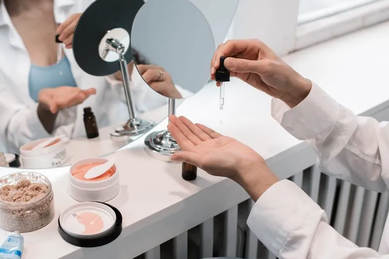 Woman letting a few drops fall into her palm at a mirrored dressing table, between skincare pots and a small brown bottle