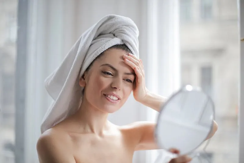 Woman with her hair wrapped in a white towel checking her skin in a hand mirror