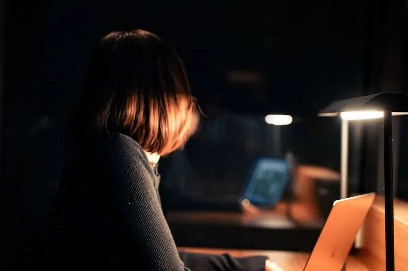 Woman seen from behind, alone at a laptop in a dark office lit by a single desk lamp