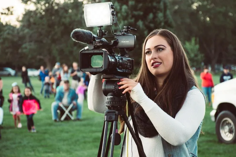 Young woman filming outdoors with a video camera on a tripod, fitted with a microphone and a light