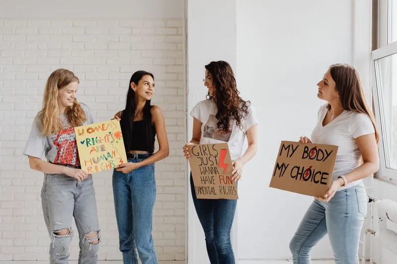 Four smiling young women against a white brick wall holding handmade signs, including 'Women's rights are human rights' and 'My body my choice'