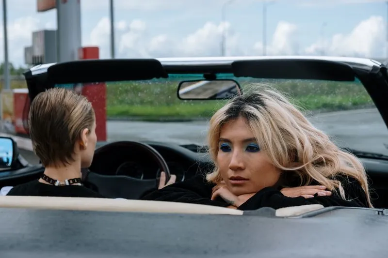 Two women in an open-top convertible, the driver seen from behind, the passenger leaning over the seat back with her chin on her arms