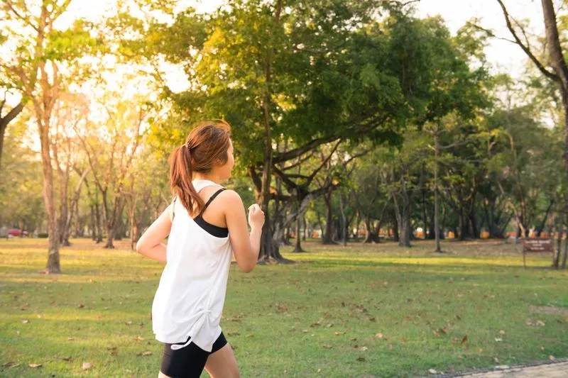 Young woman in a white vest top running through a sunlit park among tall trees