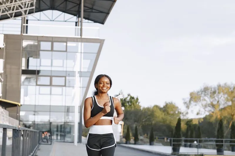 Smiling young woman in a black sports bra and shorts jogging along a promenade past a glass building in soft, low sunlight