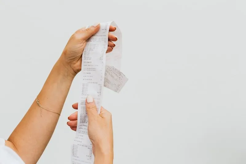 Hands holding a long, unrolled shopping receipt against a white wall