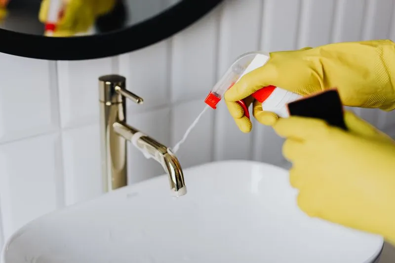 Yellow rubber-gloved hands spraying a gold-coloured tap above a white basin