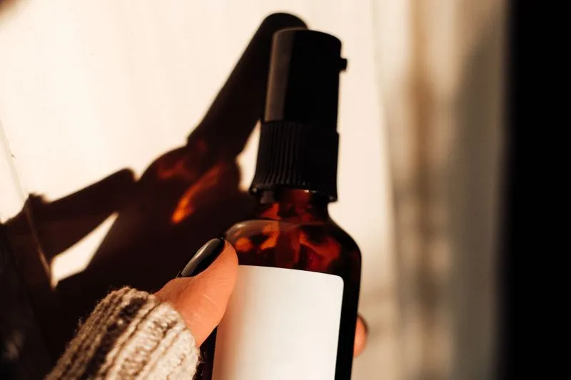 Hand holding an amber glass spray bottle with a blank white label, in low sunlight