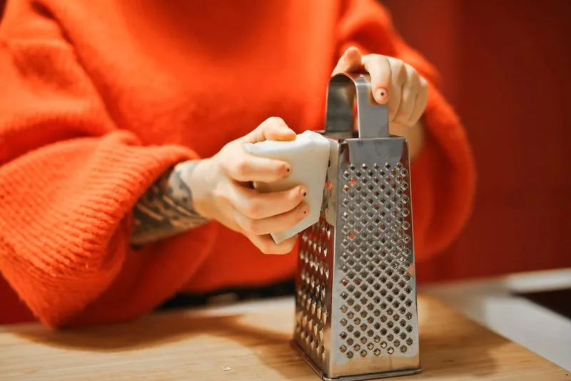 Hands grating a white bar of soap on a stainless steel box grater, on a wooden worktop