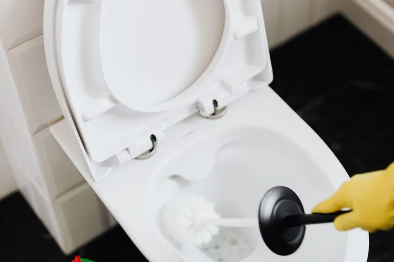 Yellow-gloved hand scrubbing the inside of a white toilet bowl with a toilet brush, seat raised