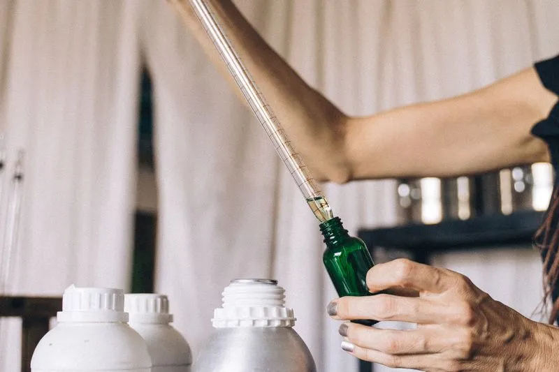 Hand measuring a liquid with a graduated pipette into a small green glass bottle, in front of white and metal canisters