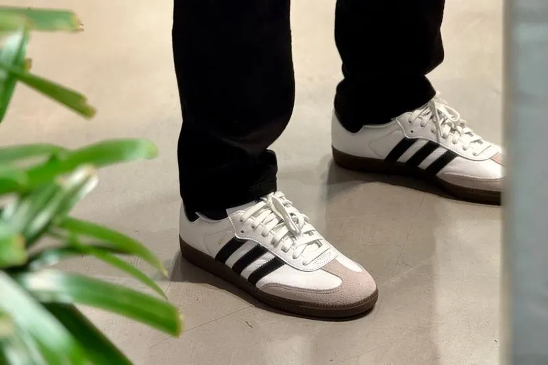 Feet in black jeans wearing white Adidas Samba trainers with black three stripes, a beige suede toe and a gum sole