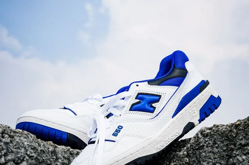 Pair of white and royal blue leather New Balance 550 trainers resting on a rock under a cloudy sky