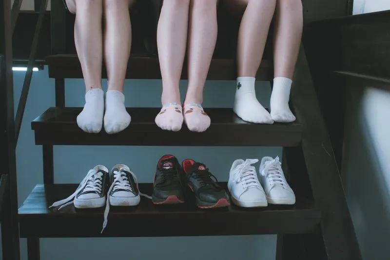 Three pairs of trainers lined up on a dark wooden stair, below the sock-clad feet of three people sitting above
