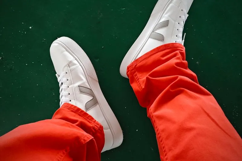 Feet in white leather Veja trainers with a grey V, worn with red trousers on a green floor