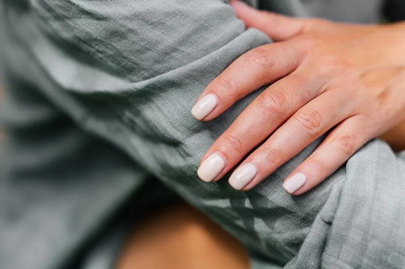 Hand with short, rounded nails in a milky nude polish, resting on sage-green linen