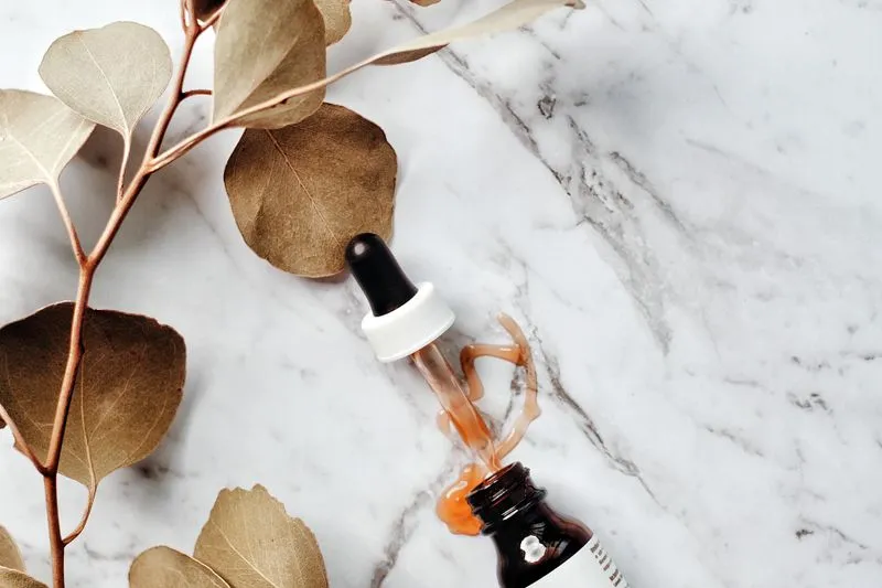 Amber serum bottle and its dropper on a marble surface, beside a dried eucalyptus sprig