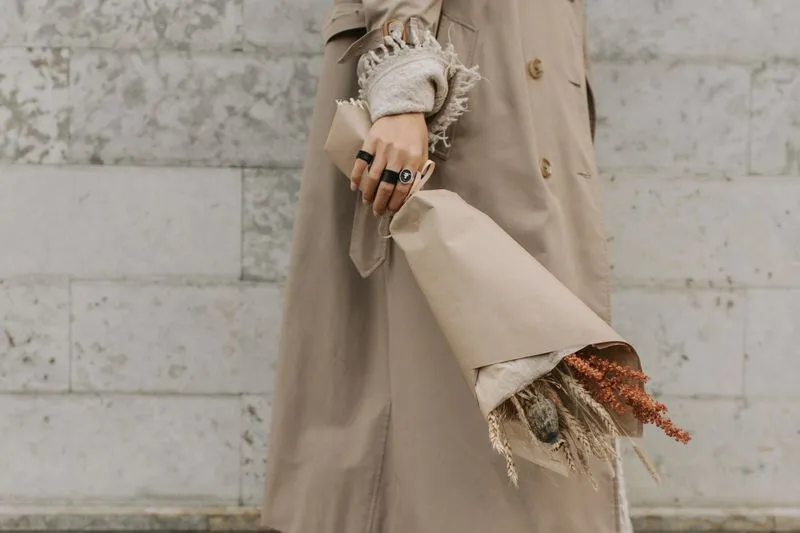 Beige trench coat worn over a cream knit, a ringed hand holding dried flowers wrapped in kraft paper