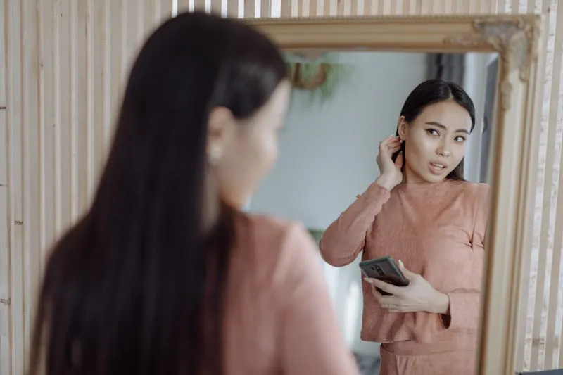 Young woman adjusting her earring in front of a mirror, phone in hand