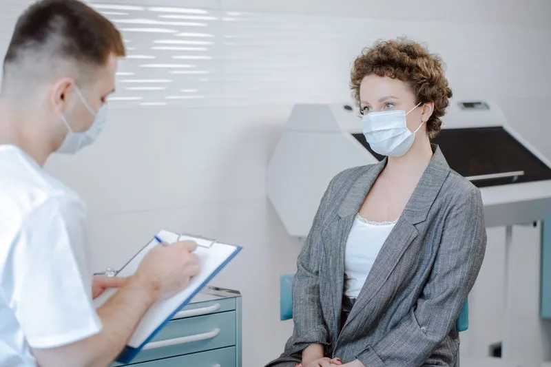 Woman patient seated opposite a doctor taking notes on a clipboard, both wearing masks