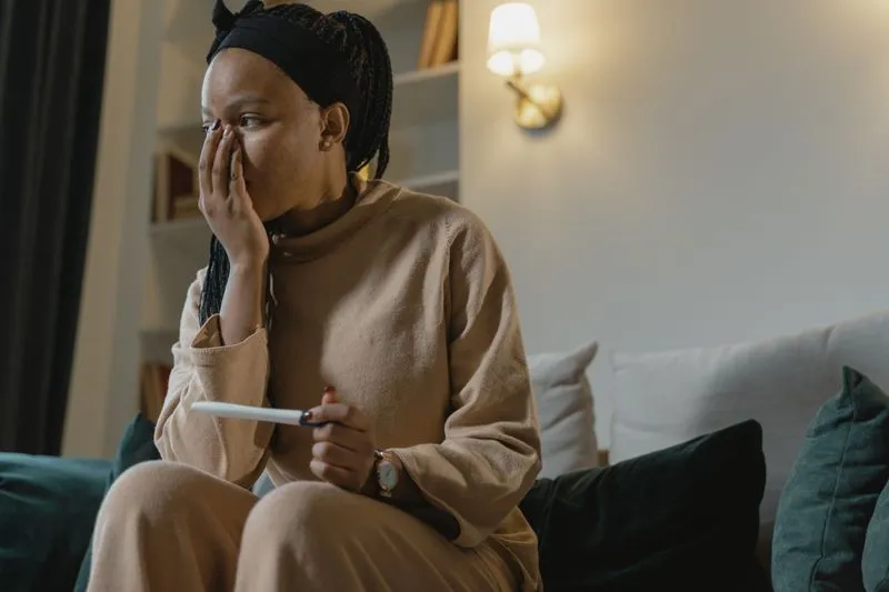 Young woman sitting on a sofa, one hand over her face and a pregnancy test in the other