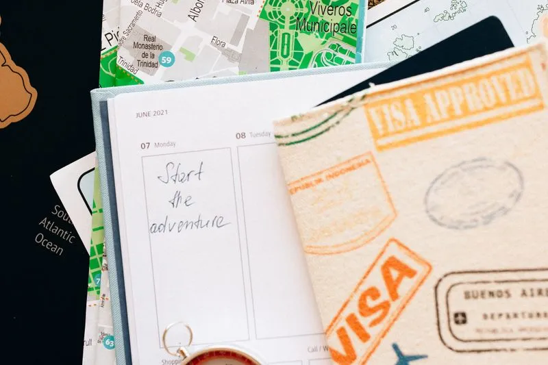 Open planner reading 'Start the adventure' on top of a city map, next to a notebook covered in visa stamps and a compass