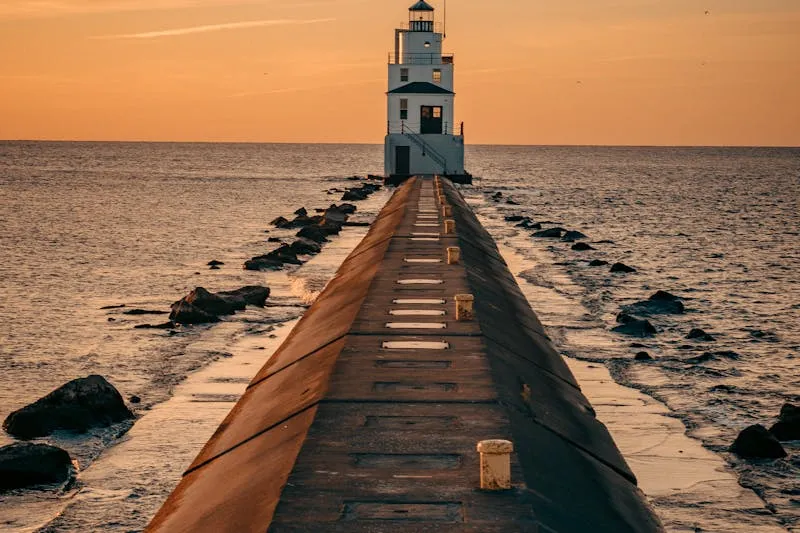 White lighthouse at the end of a long concrete pier, at sunset over calm water