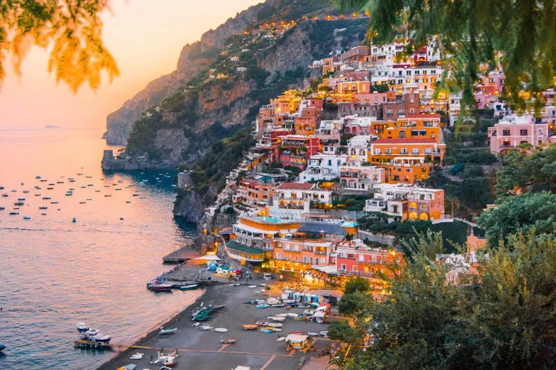 Positano at sunset seen from the hillside, colourful houses stacked above the beach and moored boats