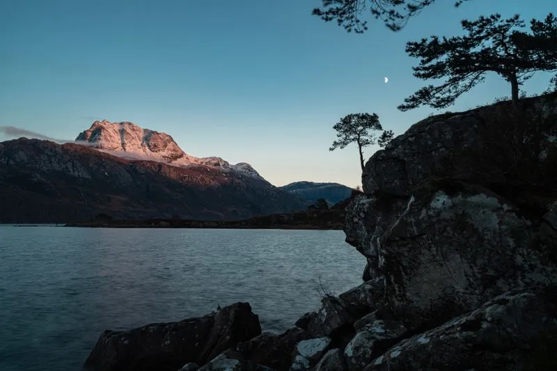 Loch and mountain in the Scottish Highlands at dusk, moon visible in the sky