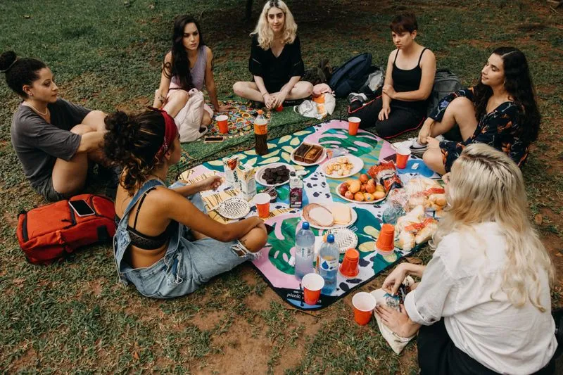 Group of friends sitting around a shared picnic on the grass, backpacks nearby