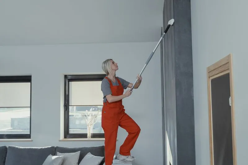 Woman in overalls standing on a low step, cleaning the top of a wall with a telescopic-handled mop
