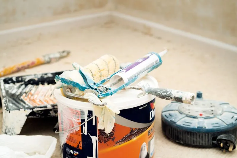 Opened tin of paint with a roller, a sealant gun and gloves on the lid, a paint tray and an extension reel on the floor