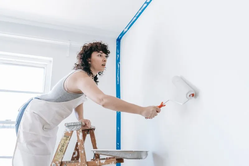 Woman leaning on a stepladder, rolling paint onto a white wall, the corner protected with blue masking tape