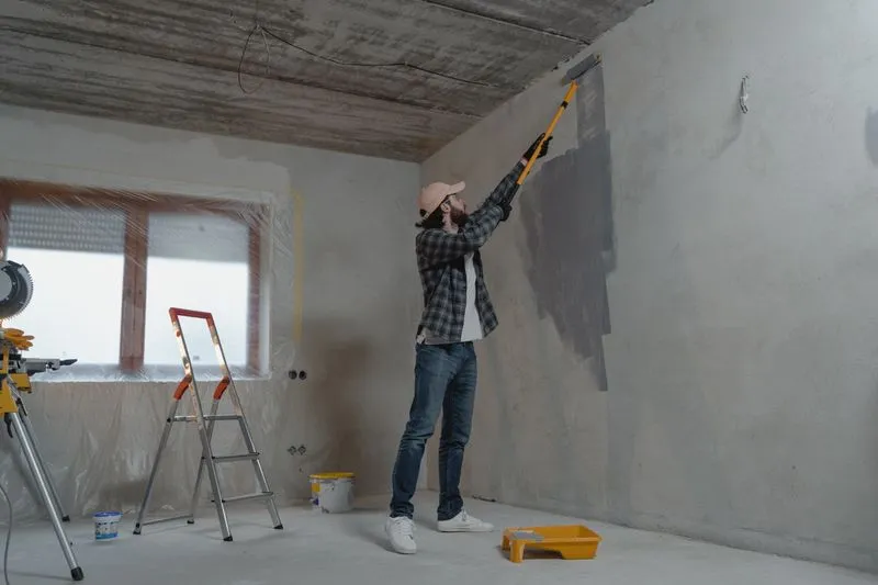 Emptied room mid-renovation, window fully covered with plastic sheeting, man rolling paint at the top of the wall with an extension pole