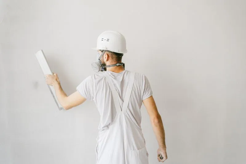 Tradesman in white overalls, hard hat and respirator smoothing a white wall with a wide filling blade