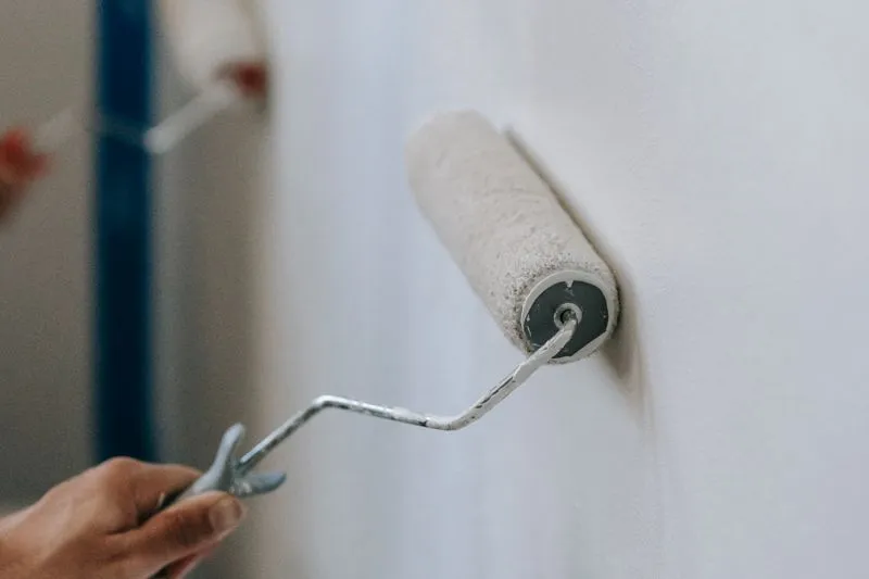 Hand running a paint roller over a white wall, blue masking tape along the corner