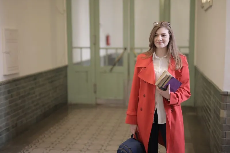 A student in a red coat holding books and a rucksack in a university corridor