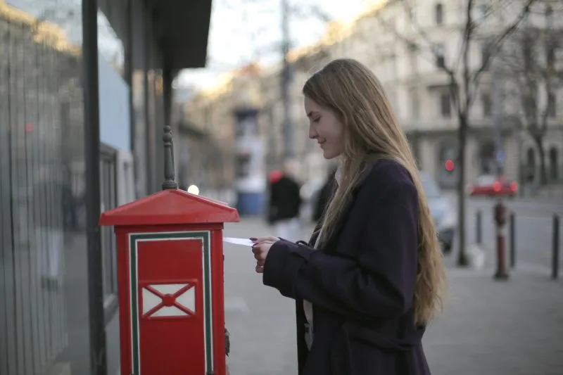 Young woman in a dark coat posting a letter into a red postbox on a city street at dusk