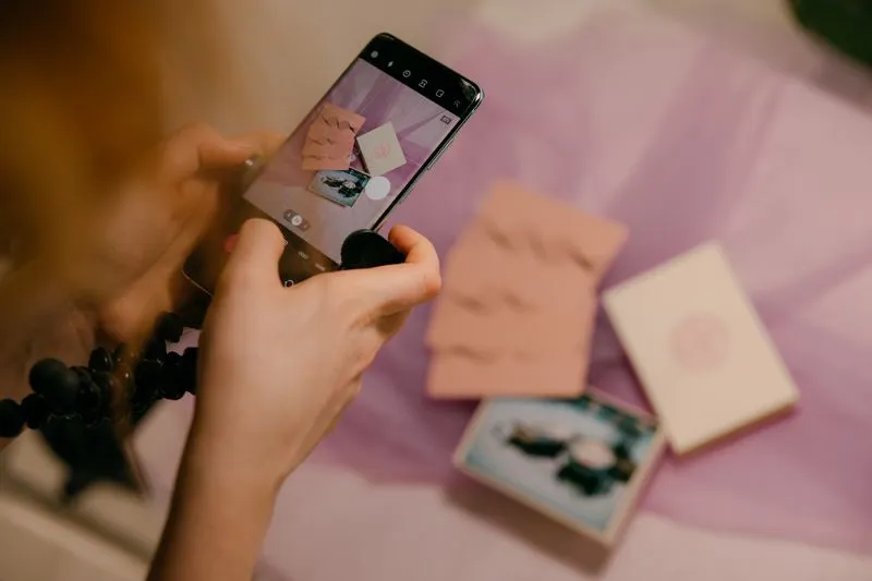 Hands photographing pink cards and envelopes with a smartphone, laid out on a lilac cloth