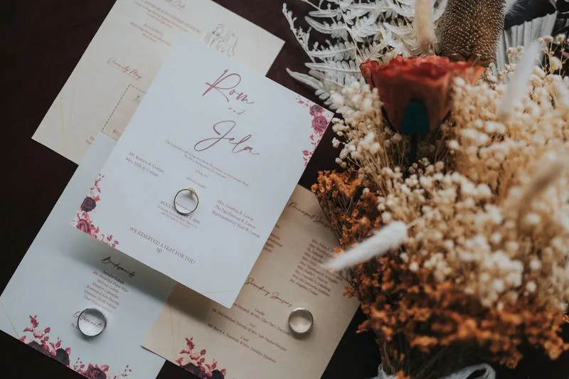 Wedding invitation and matching cards with rings laid on top, beside a bouquet of dried flowers