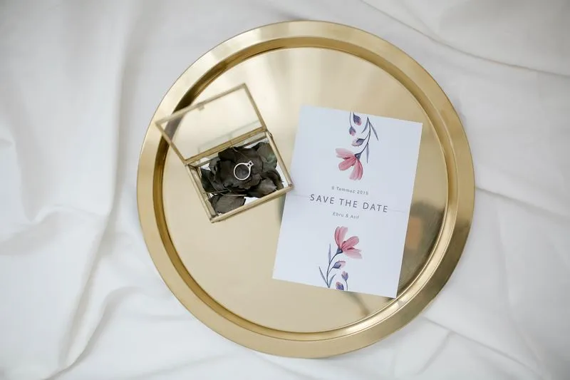 Floral save the date card on a gold tray, beside a ring in a glass box