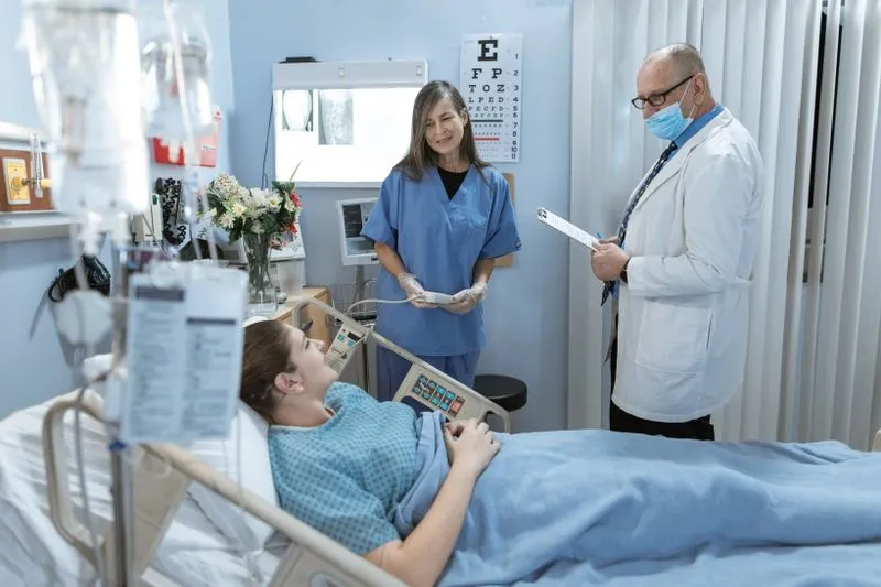 A young woman lying in a hospital bed, facing a nurse and a doctor reading her notes