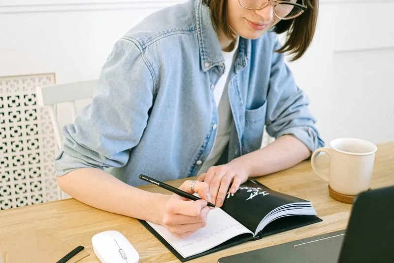 A young woman writing in an open notebook at a wooden desk next to a mug